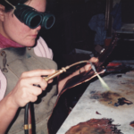 Pregnant Cathy Callihan with goggles and a torch, creating metal art, 1988